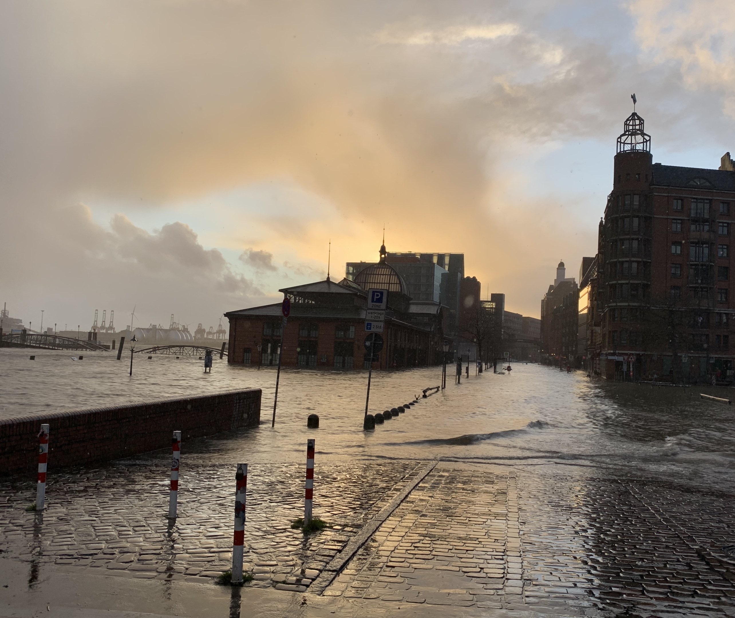 Überflutete Straße in Hamburg, Deutschland mit Wasser bedeckter Straße, Pfosten, Schilder, Gebäuden und einer Brücke unter einem bewölkten Himmel.