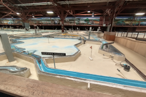 Ein großes Indoor-Wasserpark mit zahlreichen Wasserrutschen, Stangen, Texttafeln, Deckenleuchten und Wandgemälden, das wie ein Minigolfplatz gestaltet ist.
