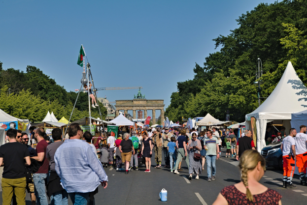 Eine Menschenmenge geht eine Straße mit Zelten, Fahrzeugen und Bäumen entlang auf einen Bogen unter einem klaren blauen Himmel zu, mit Fahnenmasten auf der linken Seite, was auf das Oktoberfest in München, Deutschland, hindeutet.
