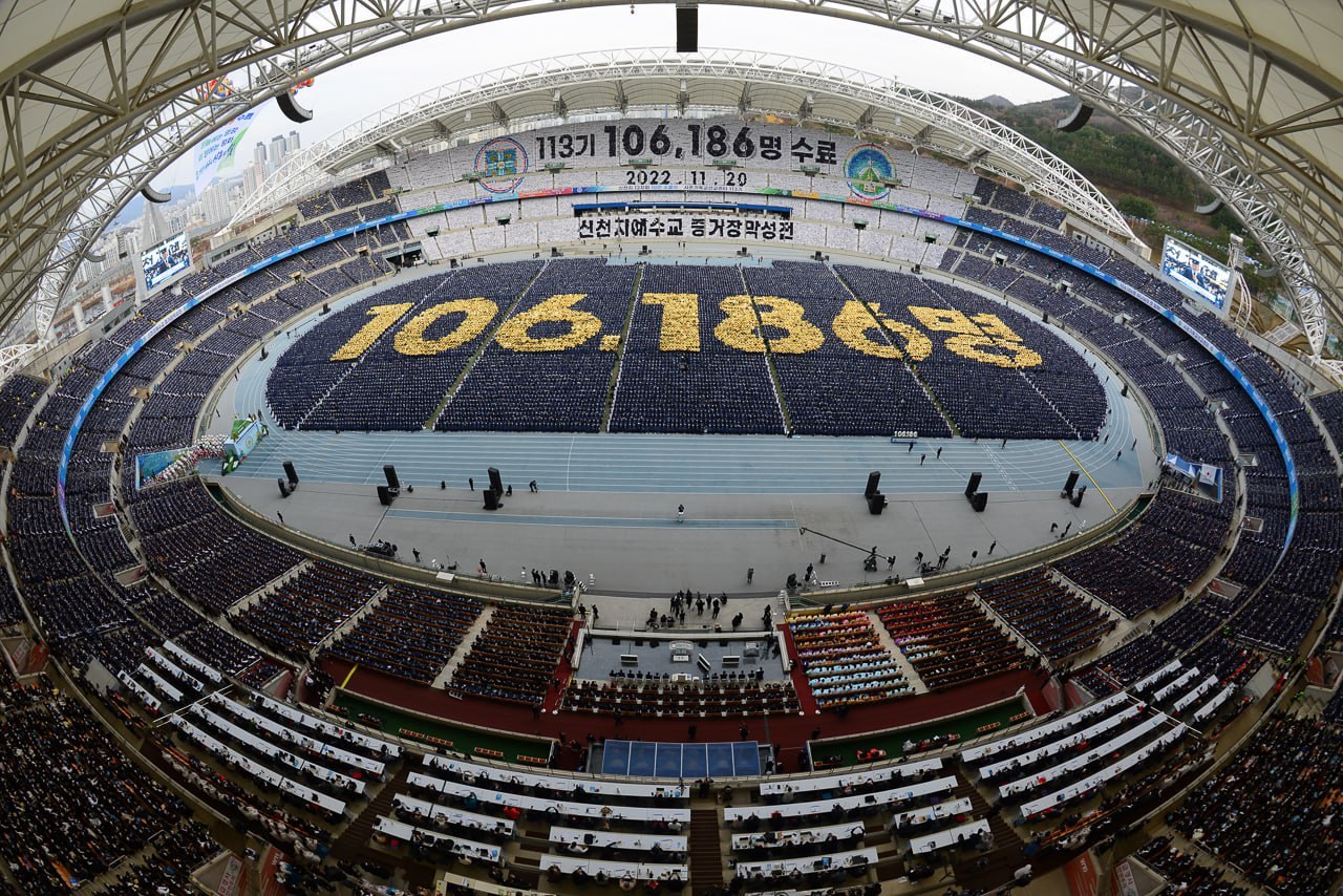 Luftaufnahme des Seoul Olympic Stadium mit Zuschauern, umgeben von Bäumen, Gebäuden und einem klaren blauen Himmel.