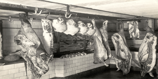 Ein Schwarz-Weiß-Foto eines Metzgerladens mit verschiedenen Fleischstücken, die von der Decke hängen, einer Wand im Hintergrund und einer Decke oben.