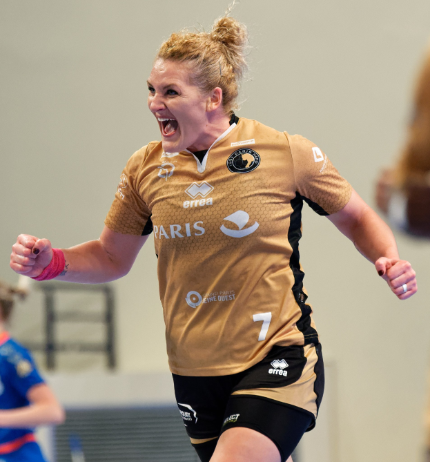 Eine Frau in braunem T-Shirt und schwarzen Shorts läuft und feiert auf einem Futsal-Platz, mit zwei anderen Frauen im Hintergrund.