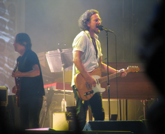 Ein Live-Musikauftritt auf der Buhne mit einem Mann in einem weissen Hemd, der in ein Mikrofon singt und Gitarre spielt, begleitet von einem anderen Mann in einem schwarzen Hemd, der Gitarre spielt, mit einem Klavier, Kabeln, Lautsprechern und Bühnenlicht im Hintergrund.