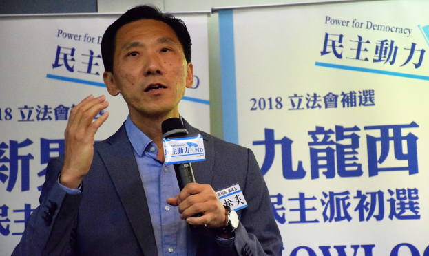 Ein Mann im Anzug spricht in ein Mikrofon mit Schildern und einer Wand im Hintergrund, trägt eine Uhr.