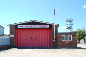 Feuerwehr- und Rettungsdienstgebäude mit roter Tür, Fenstern, einem Namensschild, einem Fahnenmast mit Flagge, einem Metallturm, einem Zaun, umgeben von Bäumen und einem bewölkten Himmel.