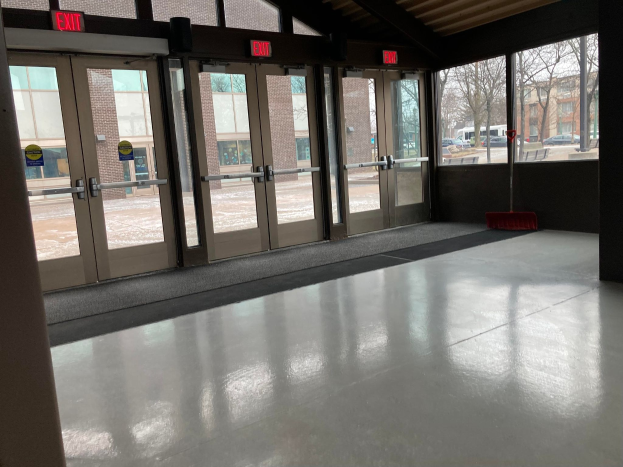 Großer Raum mit hohen Fenstern und Türen, ein Besen auf dem Boden, mehrere Schilder, ein sichtbares Dach, umliegende Bäume, geparkte Fahrzeuge, benachbarte Gebäude mit Fenstern und ein bewölkter Himmel, was auf einen kommerziellen Bodenreinigungsdienst hinweist.