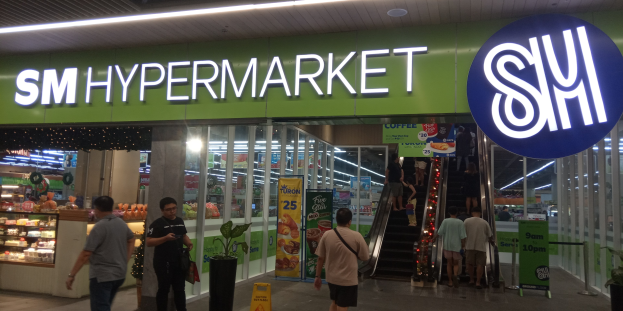 Eine Gruppe von Menschen vor einem hell erleuchteten "SM Hypermarket"-Laden mit Deckenleuchten, Texttafeln und Topfpflanzen im Inneren.