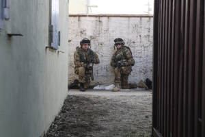 Eine Szene mit einer Wand und einem Fenster links, einer Eisenwand rechts, zwei Soldaten und Erde in der Mitte und Gebäuden, einem Strommast und einer weiteren Wand im Hintergrund.
