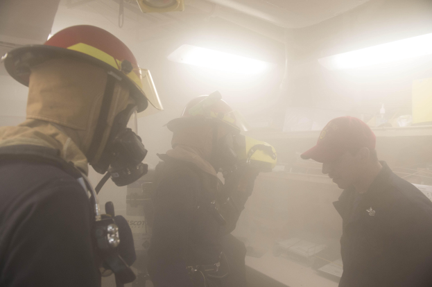 Feuerwehrleute mit Helmen und Rauch vor dem Mund, einer mit einer Kamera, während einer Trainingsrunde mit Hintergrundbeleuchtung und -ausrüstung.