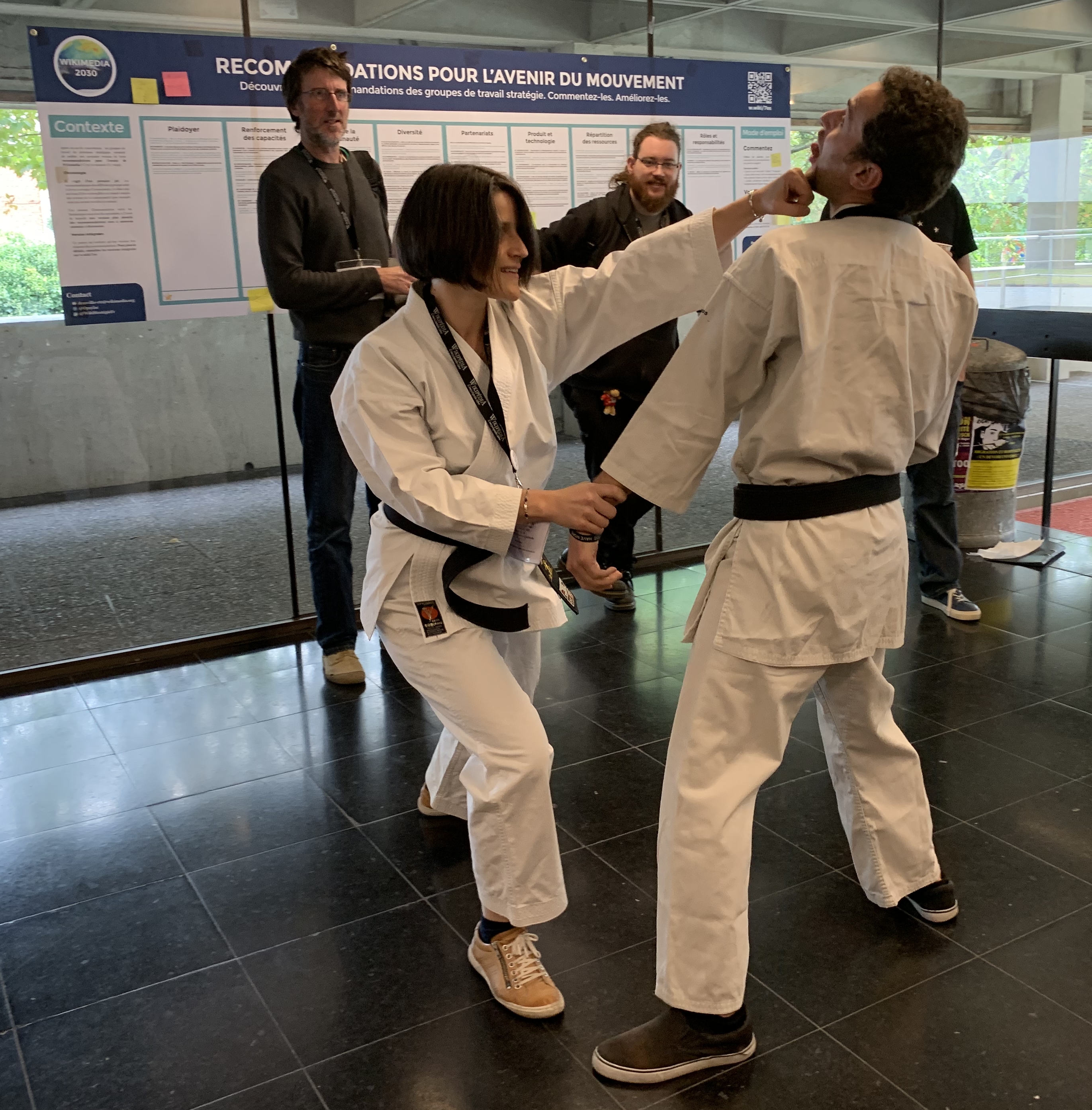 Ein Mann und eine Frau in Karate-Ausrüstung stehen nebeneinander vor einem Plakat, mit einer Glaswand, die Bäume dahinter zeigt, und einem Mülleimer und einem Tisch mit Gegenständen rechts daneben.