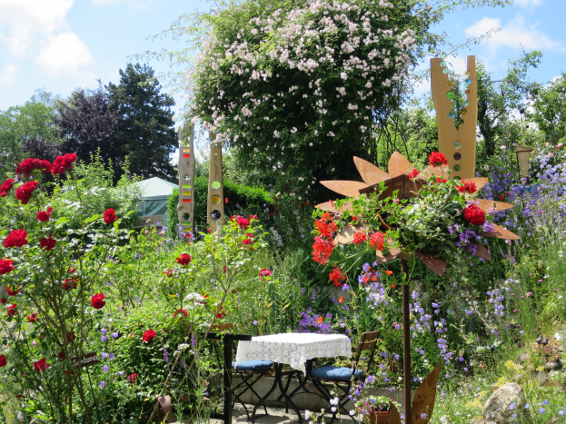 Ein Garten auf der Chelsea Flower Show mit bunten Blumen und Pflanzen, einem Tisch mit einer Decke umgeben von Stühlen und Bäumen unter einem bewölkten Himmel.