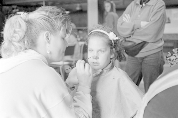 Ein junges Mädchen lässt sich von einer Frau in einem Salon Make-up auftragen, während zwei andere Personen im Hintergrund stehen, alles in Schwarz-Weiß.
