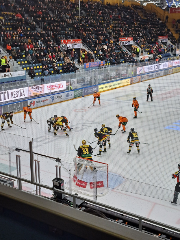 Eine Gruppe von Menschen, die Hockey auf einer Indoor-Eisfläche spielen, umgeben von einem Zaun, mit Zuschauern im Hintergrund und beleuchteten Stadionlampen oben.