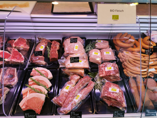 Eine Vitrine in einer Metzgerei, die verschiedene Fleisch- und Wurstsorten in Schalen mit Preisschildern präsentiert, mit einem Textschild im Hintergrund.