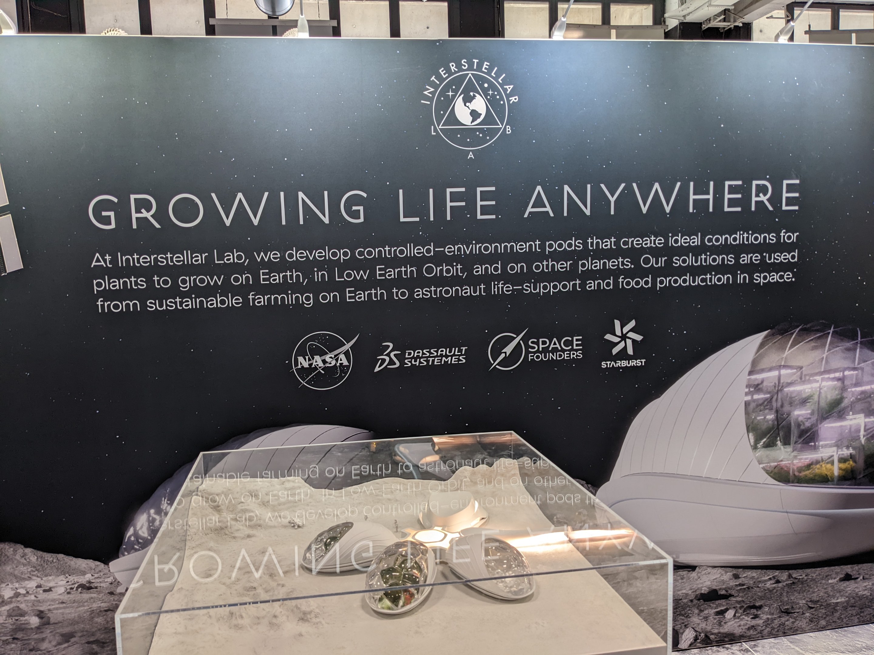 Stand mit einer "Wachse Leben Überall"-Tafel, der einen Tisch mit Gegenständen im Vordergrund und eine Displaytafel mit Text und Bildern im Hintergrund zeigt, Teil der Interstellar Lab-Ausstellung.