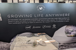 Stand mit einer "Wachse Leben Überall"-Tafel, der einen Tisch mit Gegenständen im Vordergrund und eine Displaytafel mit Text und Bildern im Hintergrund zeigt, Teil der Interstellar Lab-Ausstellung.