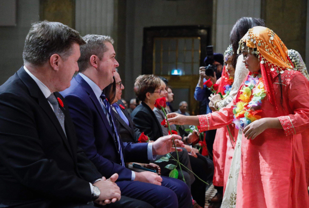 Eine Gruppe von Menschen, die in einem Raum zusammensitzen, mit einer Frau in der Mitte, die einen Kranz trägt und Blumen hält, flankiert von einem Mann im Anzug und einer Person mit einer Kamera, vor einer Wand mit einer Tür.