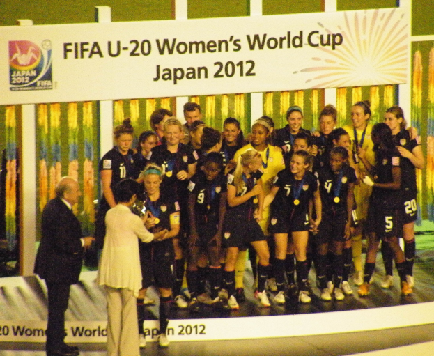 Gruppe von Frauenfußballspielerinnen in schwarzen Uniformen, die eine Trophäe auf einer Bühne halten, mit einer Tafel im Hintergrund.