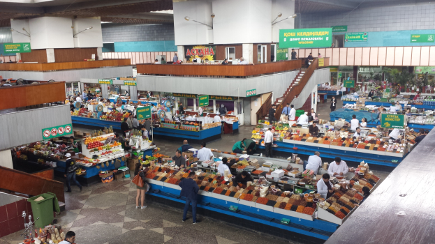 Großer Markt mit zahlreichen Menschen, Tischen mit Speisen, beleuchteter Decke, Treppen mit Geländern und einem Müllcontainer auf der linken Seite.