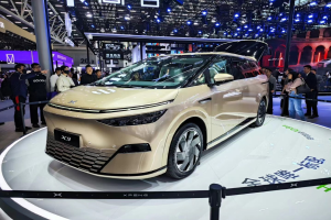 Das Xpeng X9 Auto auf einer Automesse, umgeben von Menschen, mit Pfosten, einem Zaun, einem Display-Bildschirm, einem Gebäude und einem Dach mit Deckenbeleuchtung im Hintergrund.