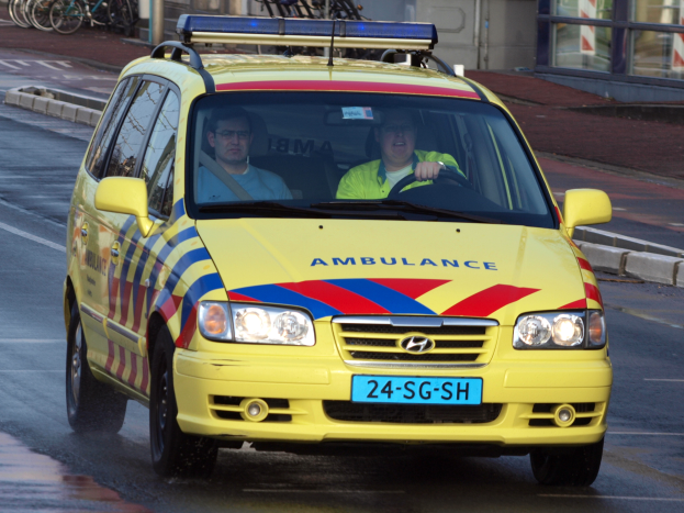 Ein gelber Krankenwagen mit zwei Insassen fährt auf einer Straße, mit parkenden Fahrrädern auf dem linken Gehweg und einem Glasfenstergebäude im Hintergrund.