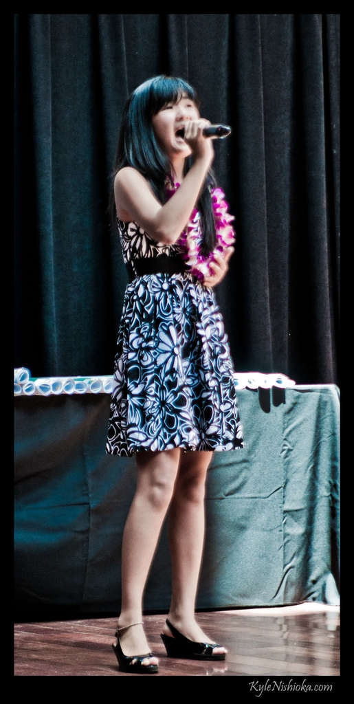 Eine Frau in einem bunten Kleid singt auf der B├╝hne in ein Mikrofon, mit einem tisch voller Papiere und einem Vorhang im Hintergrund.