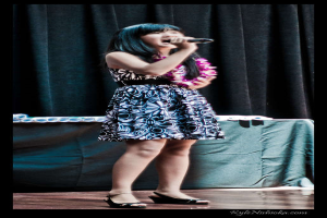 Eine Frau in einem bunten Kleid singt auf der B├╝hne in ein Mikrofon, mit einem tisch voller Papiere und einem Vorhang im Hintergrund.