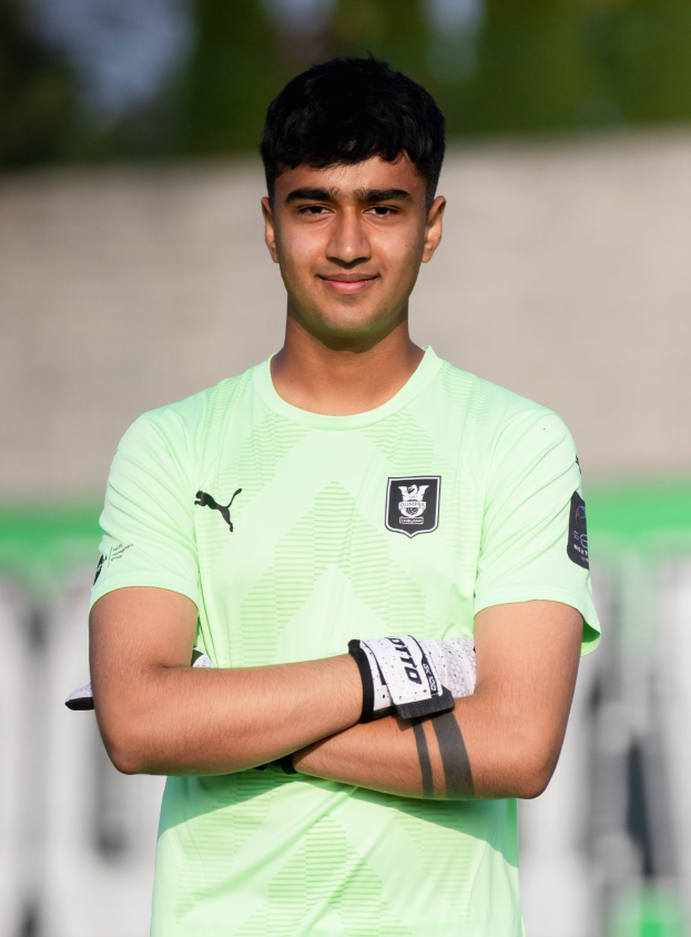 Ein junger Mann in grünem T-Shirt steht auf einem Fußtballfeld mit verschräckten Armen, lächelnd, wahrscheinlich ein Torwart, mit einem unscharfen Hintergrund.