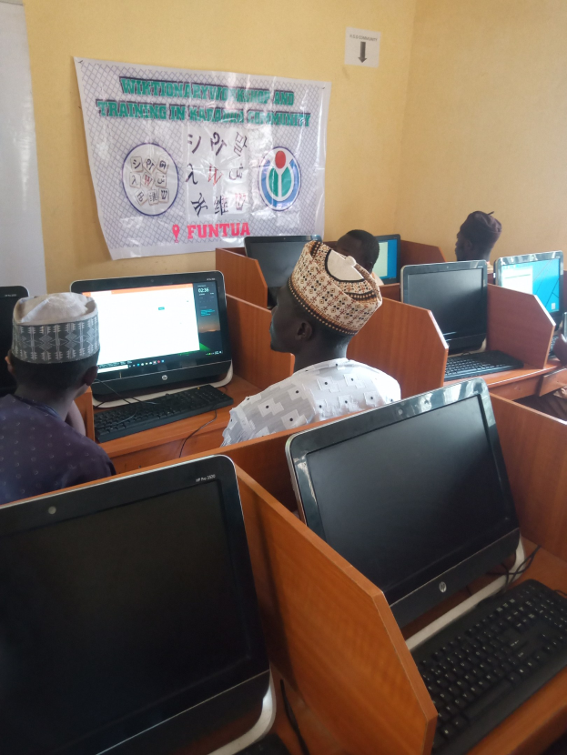 Eine Gruppe von Menschen sitzt an Tischen mit Computerbildschirmen, Tastaturen und Mäusen und nimmt an einem Workshop zu Computertechnologie teil, wobei ein Plakat im Hintergrund zu sehen ist.