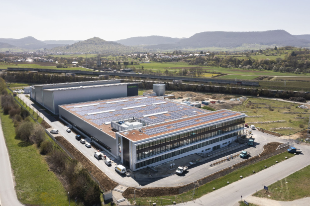 Luftaufnahme eines großen Gebäudes mit Solarmodulen auf dem Dach, umgeben von Fahrzeugen, Bäumen, Gras und Masten, vor einem Hügel- und einem klaren blauen Himmel.