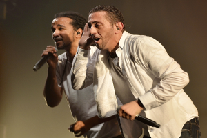 Zwei Männer in legerer Kleidung treten nebeneinander auf der Bühne auf, jeder hält ein Mikrofon in der Hand.