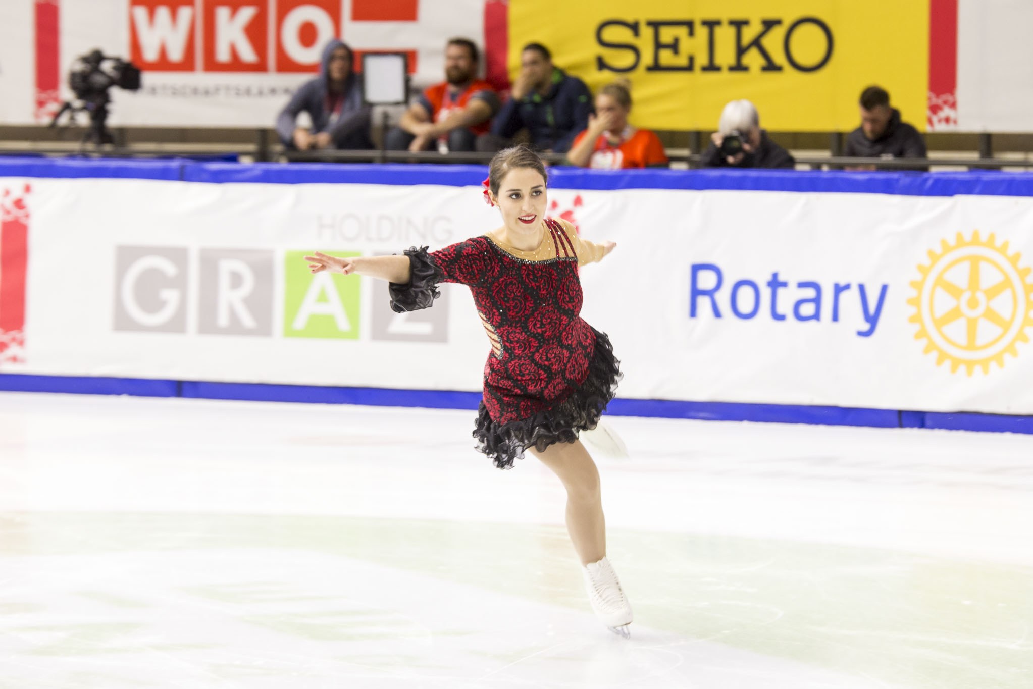 Eine Frau in einem roten und schwarzen Kleid, die auf einer Eisbahn während der Weltmeisterschaft für Eiskunstlauf skatet, mit Zuschauern und einem Fotografen im Hintergrund.