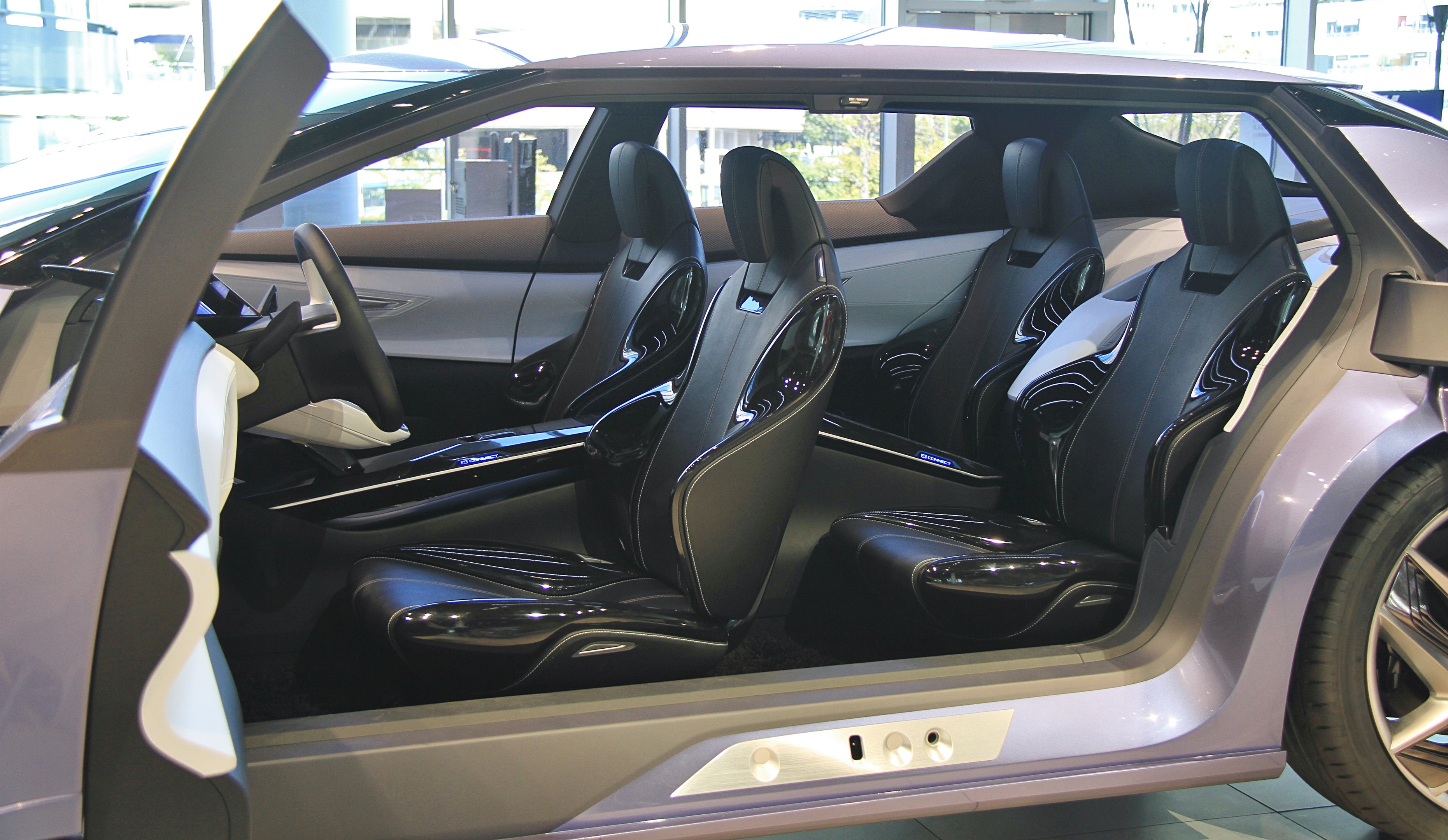 Innenansicht eines Autos mit offener Tür, die lederne Sitze, Lenkrad und Rad zeigt, mit Gebäuden und Bäumen im Hintergrund.