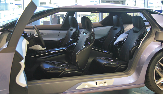 Innenansicht eines Autos mit offener Tür, die lederne Sitze, Lenkrad und Rad zeigt, mit Gebäuden und Bäumen im Hintergrund.