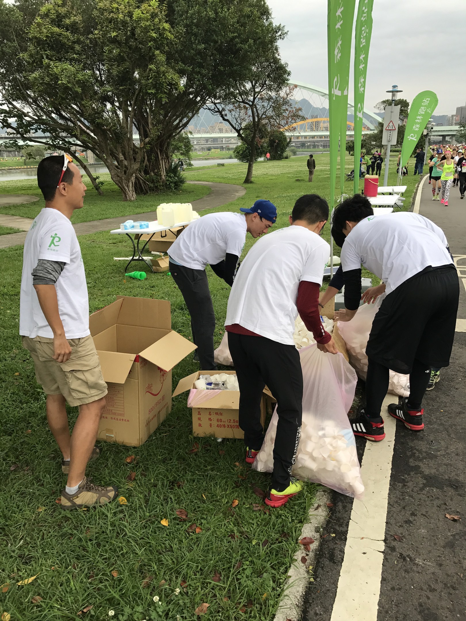 Group of people on a roadside with scattered cardboard boxes and bottles, participating in a charity run with banners, trees, buildings, and a clear blue sky in the background.