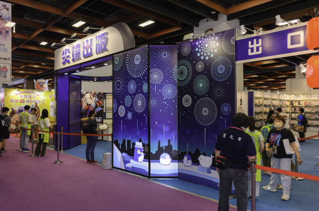 Eine Gruppe von Menschen an einem Messestand mit Absperrpollern, Bannern, Deckenleuchten und hängenden Dekorationen, einige tragen Taschen.