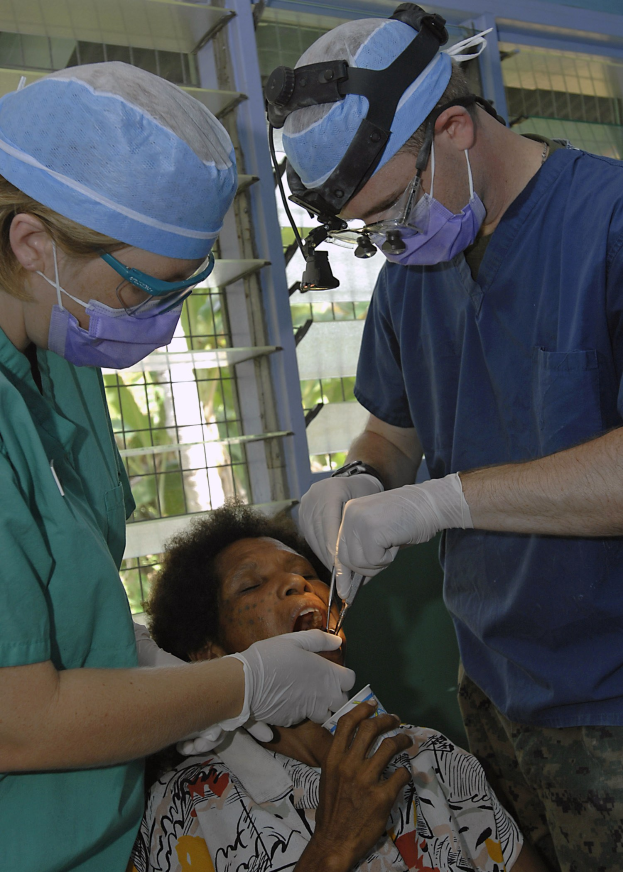 Zwei Chirurgen in Operationskleidung führen eine Gesichtsoperation an einer Frau auf einem Bett durch, mit einem Fenster und Bäumen im Hintergrund.