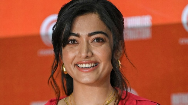 Frau in einem roten Kleid mit einem eleganten Dutt und dezentem Make-up, die in die Kamera lächelt, trägt schwarze High Heels, mit einem hellgelben Banner im Hintergrund, das 'Sonam Kapoors beste Schönheits Looks vom Cannes Film Festival' lautet.