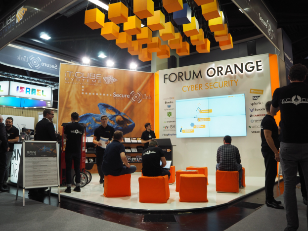 Eine Gruppe von Menschen an einem IT-Cube Systems Forum Orange Cyber Security Stand auf einer Messe, mit sitzenden und stehenden Teilnehmern, einer Tafel mit Text, einem Bildschirm, Regalen mit Gegenständen, Schildern, Deckenlampen und Lampen.