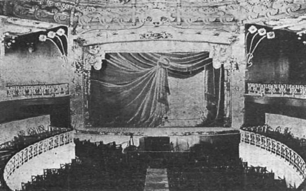 Schwarzes und weißes Foto des Auditoriums des Theatre Royal in London, das eine Bühne mit Vorhang, Geländern und ornamentalen Wanddesigns zeigt, sowie ein paar sitzende Menschen und Text am unteren Rand.