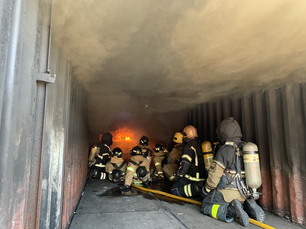 Feuerwehrleute in Schutzausrüstung mit Rauch und Feuer in einem Tunnel, einer hält ein Rohr, mit einer Wand links.