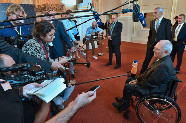 Ein Mann im Anzug im Rollstuhl spricht zu Reportern mit Kameras und Mikrofonen, mit einem Banner und einer Tür im Hintergrund.