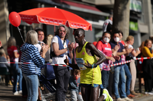 Ein Mann in einem gelben T-Shirt und Laufschuhen sprintet durch einen Marathon, während eine Menge hinter ihm jubelt, mit einem Schirm und einem Kinderwagen im Hintergrund.