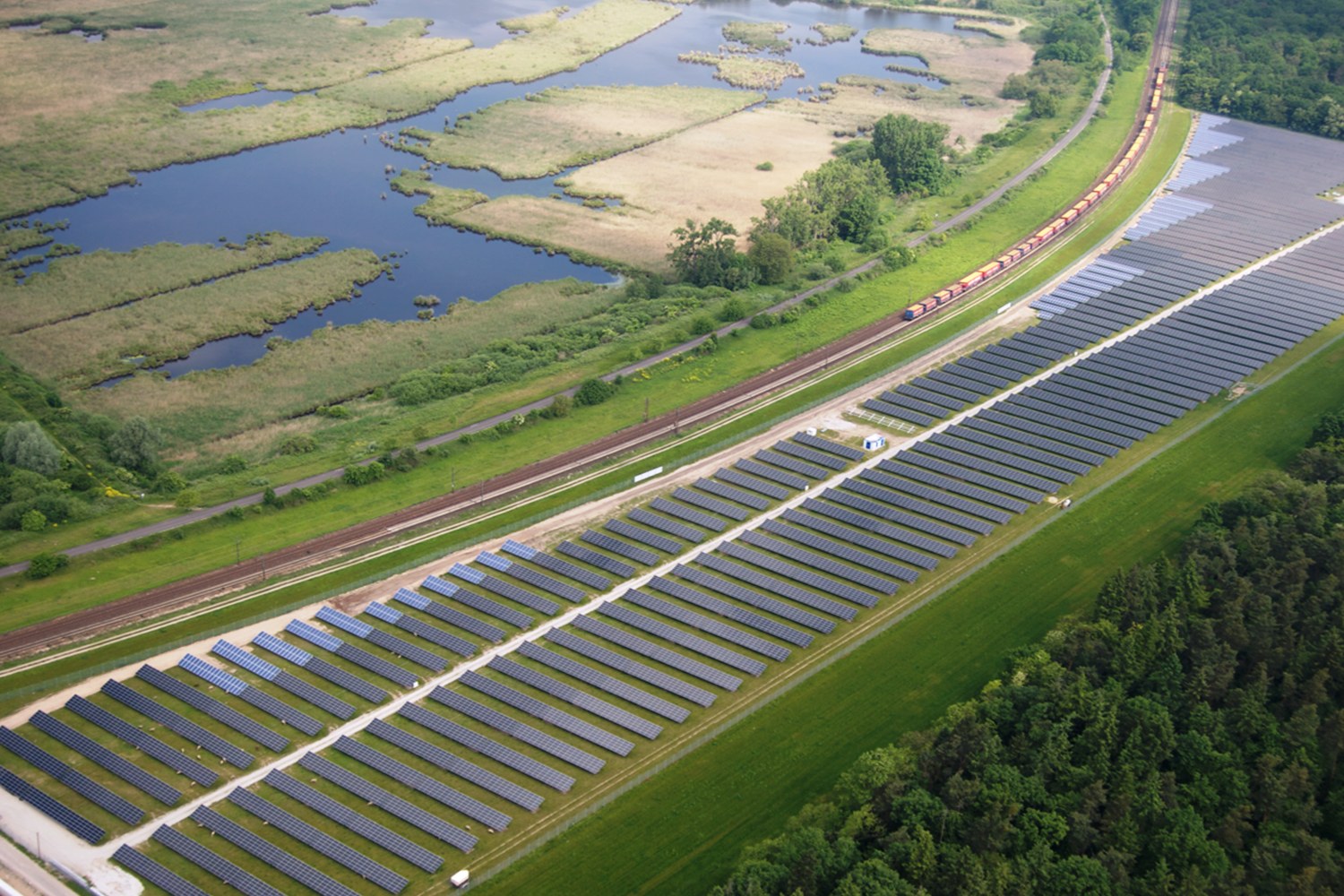 Luftaufnahme eines Solarparks mit Solarpanelen auf einem Feld, umgeben von Bäumen, Gras, Pflanzen und Wasser, mit einem Zug auf einem nähergelegenen Bahngeleis.