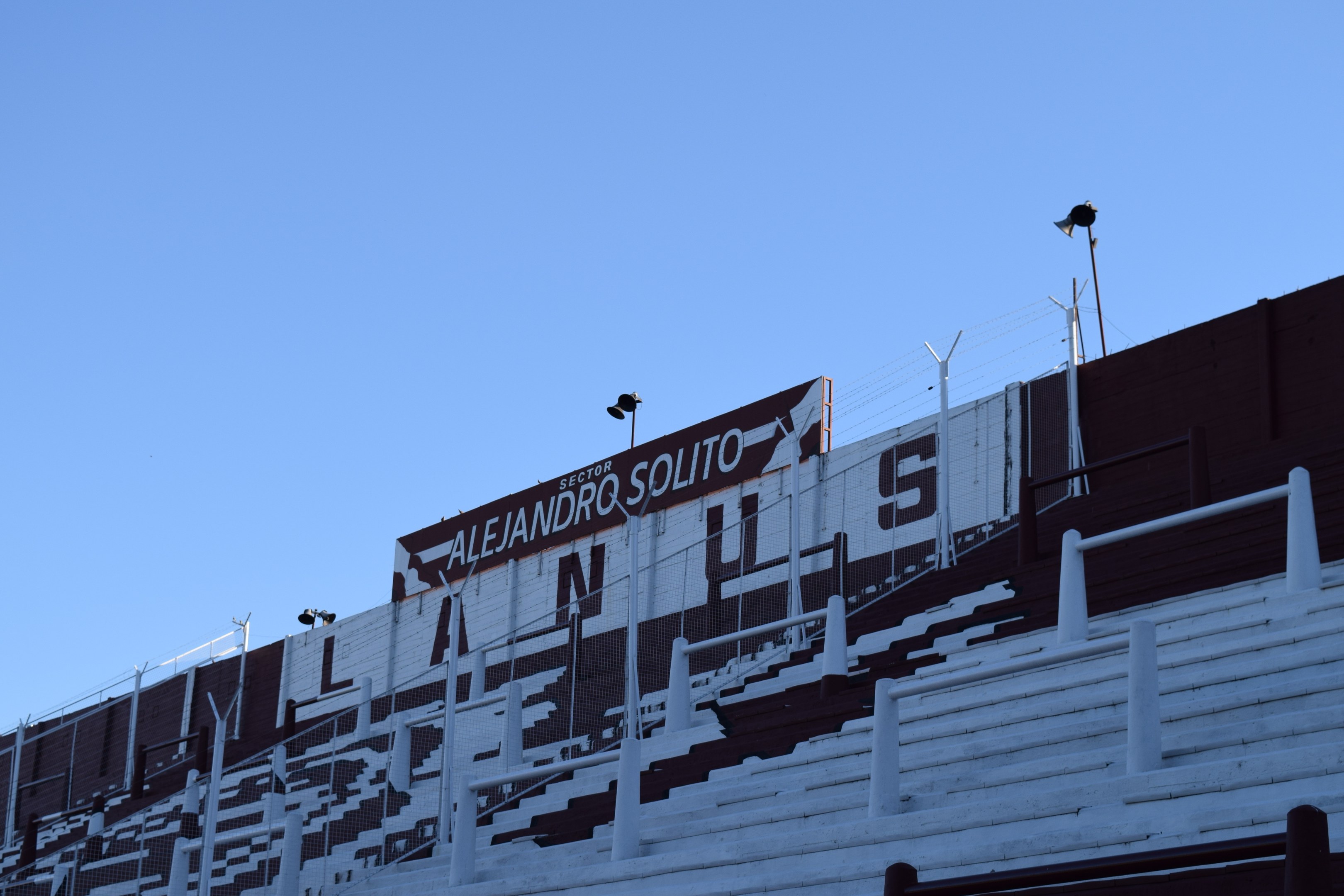 Fußballstadion mit Treppen, Geländern, Pfosten, Lampen und einer Seitentafel unter einem sichtbaren Himmel.