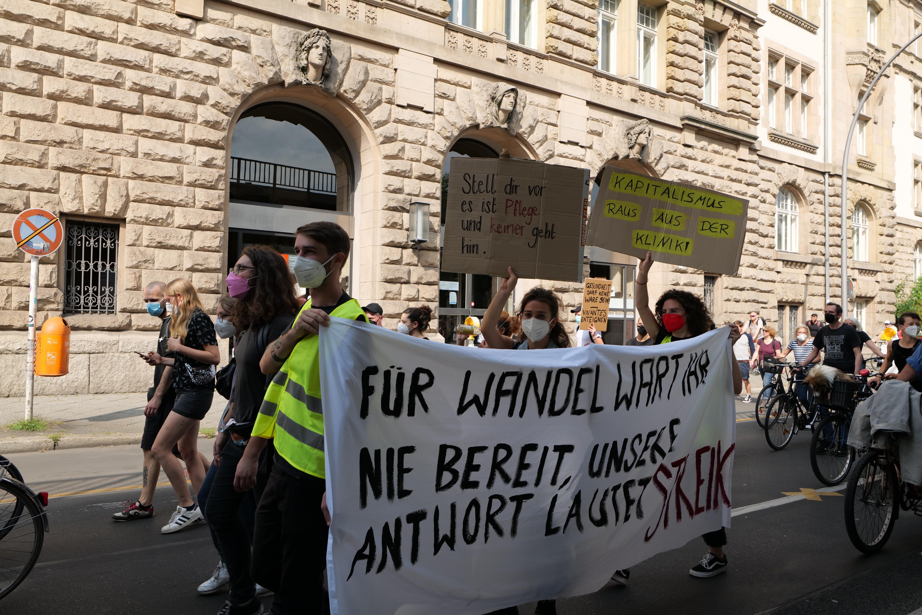 Eine Gruppe von Menschen, die die Straße entlanggehen, einige mit Plakaten und Bannern, andere mit Fahrrädern, mit einem Gebäude mit Fenstern, Bögen, Säulen und Skulpturen im Hintergrund bei einer Demonstration gegen die Anti-Kriegsbewegung in Berlin.