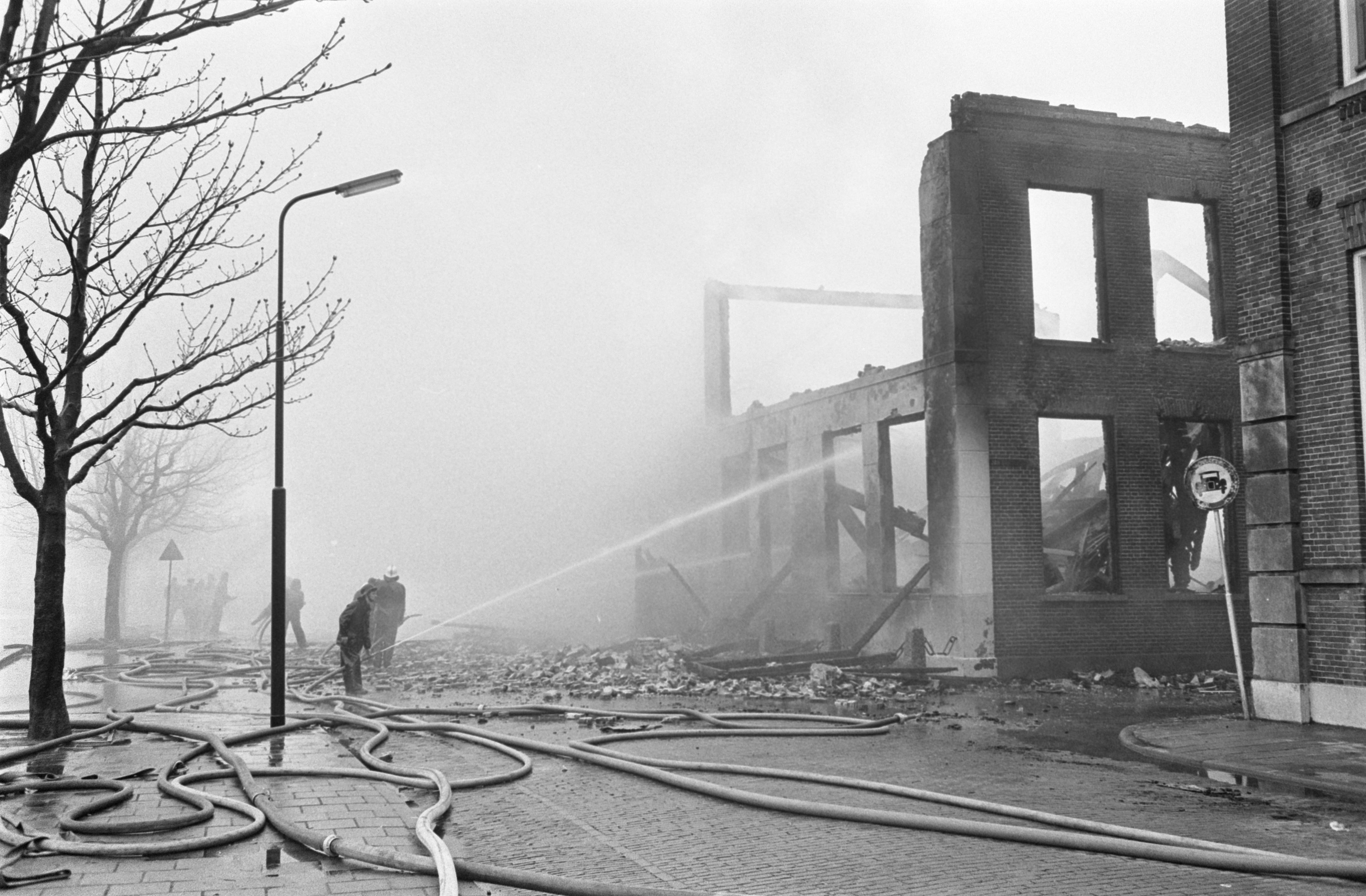 Schwarzes und weißes Bild eines feuerbeschädigten Gebäudes mit Feuerwehrleuten, die Wasser spritzen, umgeben von Menschen, Trümmern, Bäumen, einer Straßenlaterne und einem Schild unter einem bewölkten Himmel.