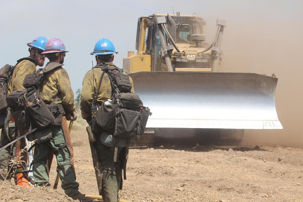 Drei Feuerwehrleute in Helmen und mit Taschen neben einem Bulldozer auf dem Boden stehend, mit Bäumen und einem klaren blauen Himmel im Hintergrund.