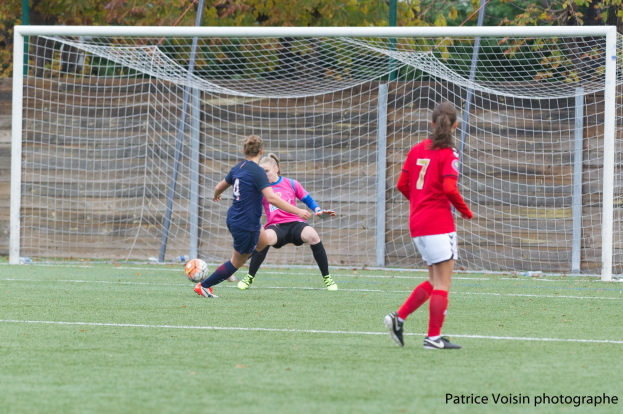 Gruppe von Frauen beim Fußballspielen auf einem Rasenfeld mit Bäumen im Hintergrund und einem Torpfosten im Bild. Text erscheint unten im Bild.