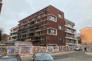 Großes Gebäude mit Gerüst an der Seite, umgeben von Bäumen und einem klaren blauen Himmel, mit Fahrzeugen auf der Straße und einer Person auf dem Bürgersteig, was auf laufende Bauarbeiten hinweist.